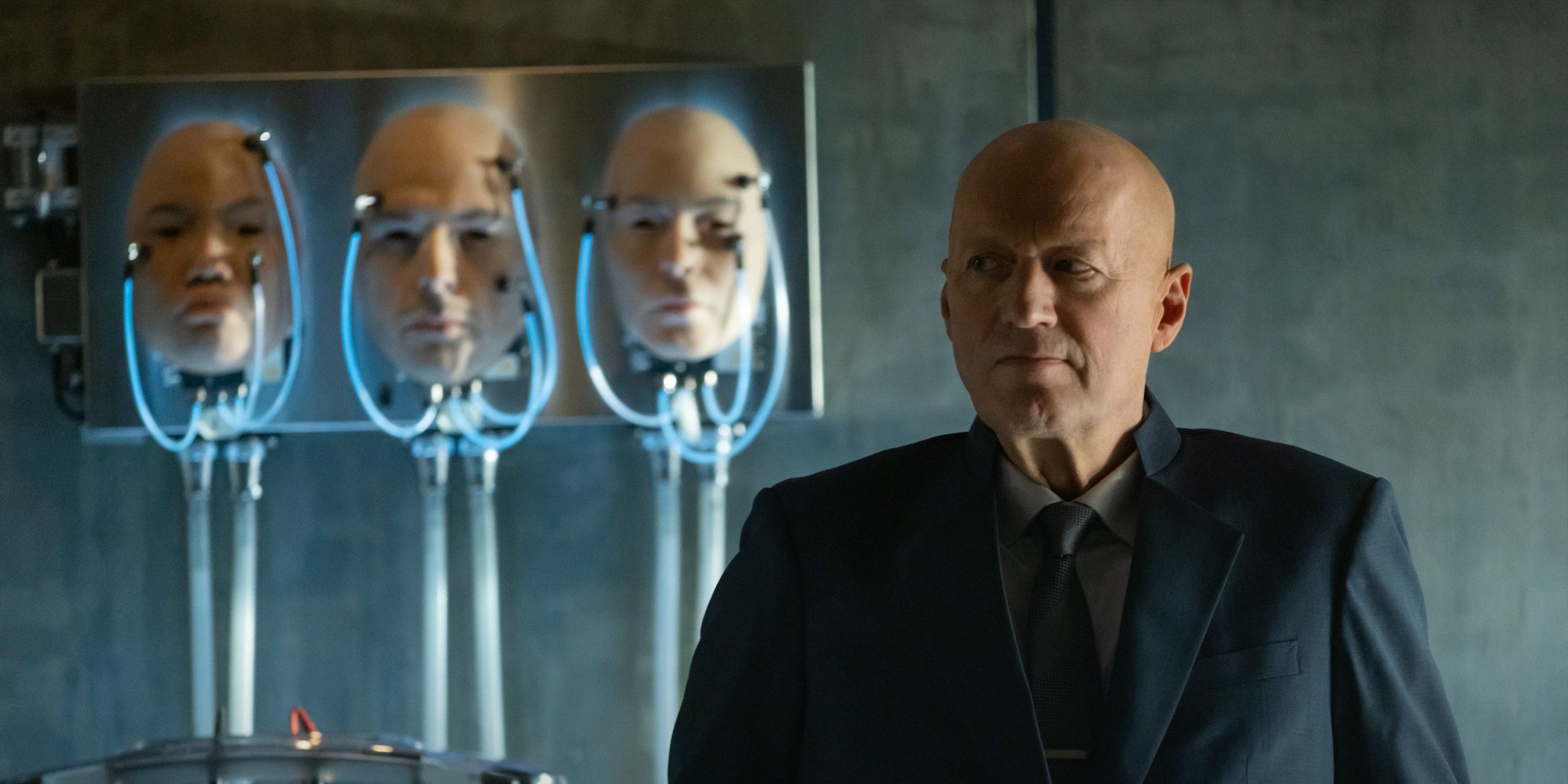 Ade Edmondson dans le rôle d'Atom Eins. debout à côté de quelques têtes prothétiques dans Alien: Earth.