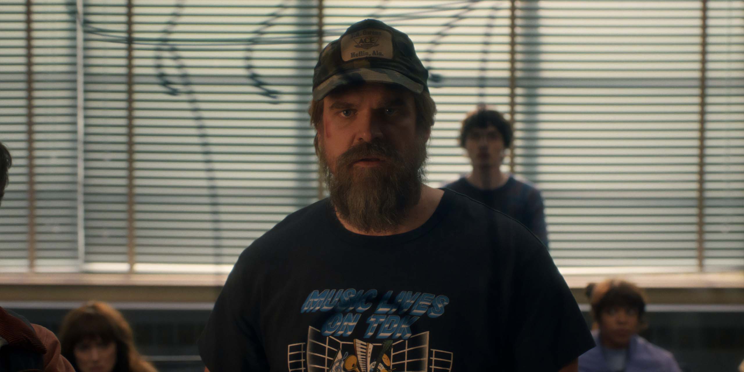 Jim Hopper, dans la saison 5 de Stranger Things, a l'air inquiet, avec Joyce, Mike et Erica en arrière-plan.