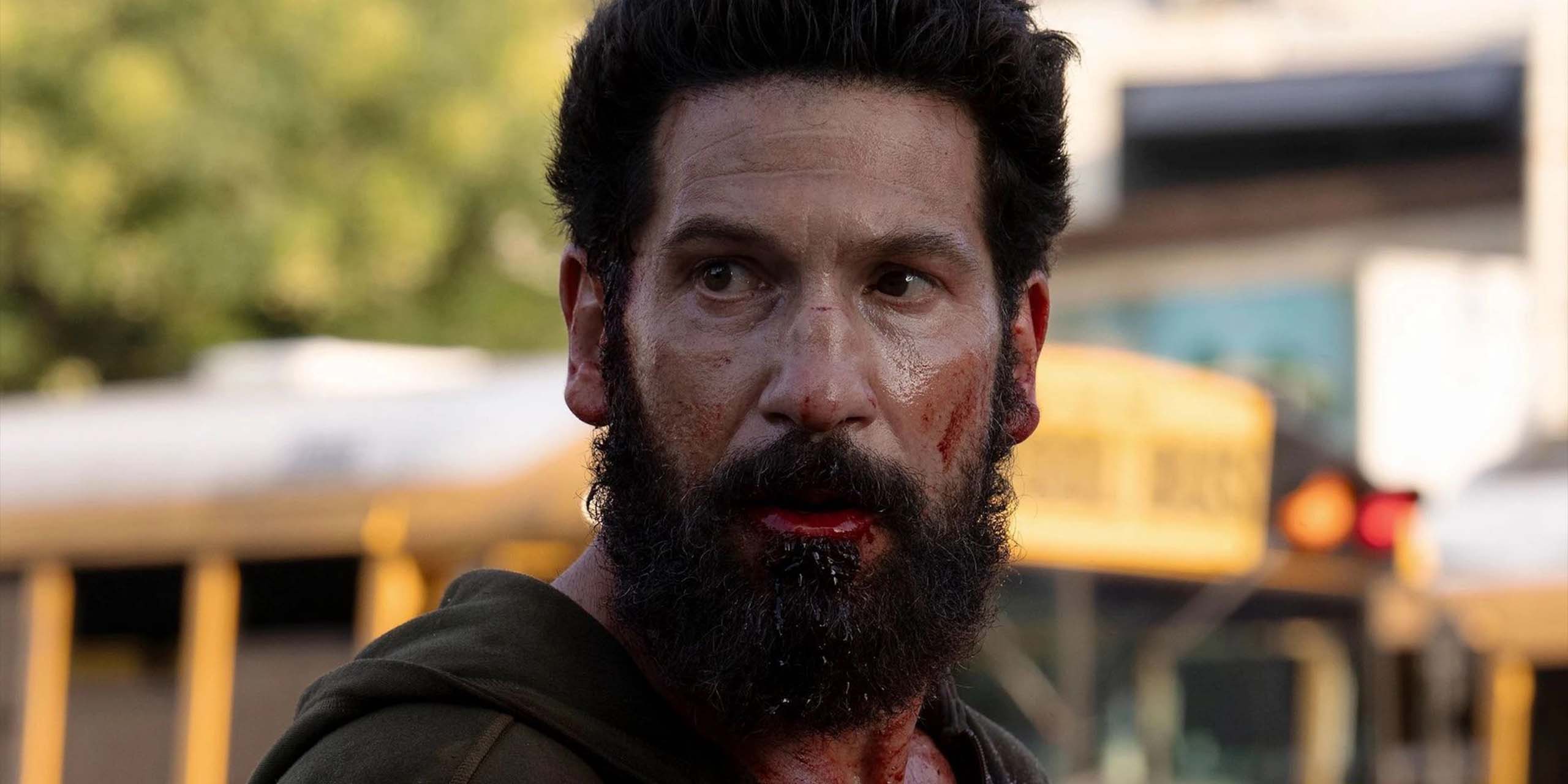 Frank Castle, barbu, regarde quelque chose en passant devant des bus scolaires dans The Punisher : One Last Kill