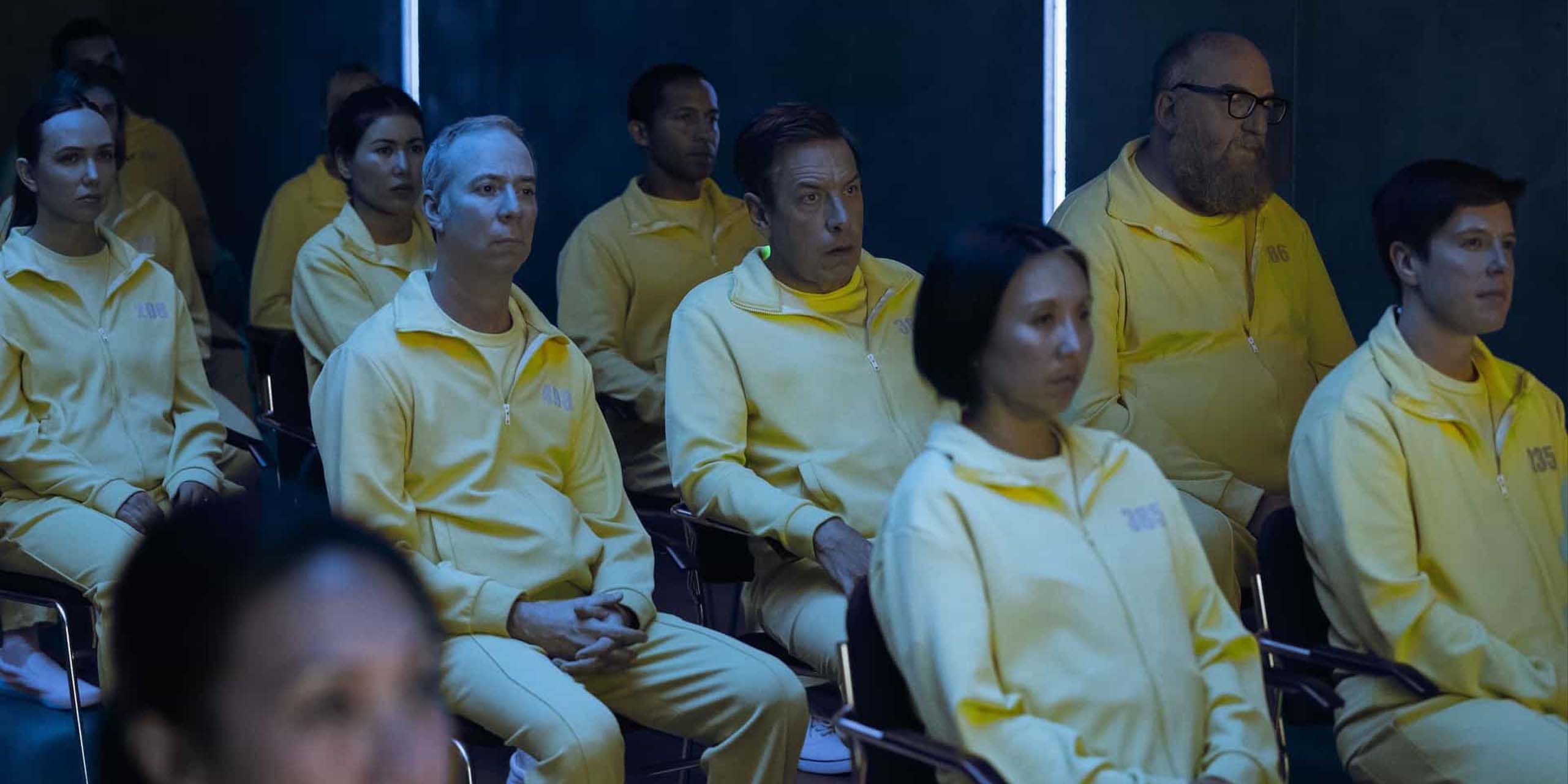 Kevin Sussman, John Ross Bowie, et Brian Posehn en prison, assis dans une salle face à une vidéo de propagande, habillés d'une combinaison jaune dans la saison 1 de Stuart Fails to Save the Universe