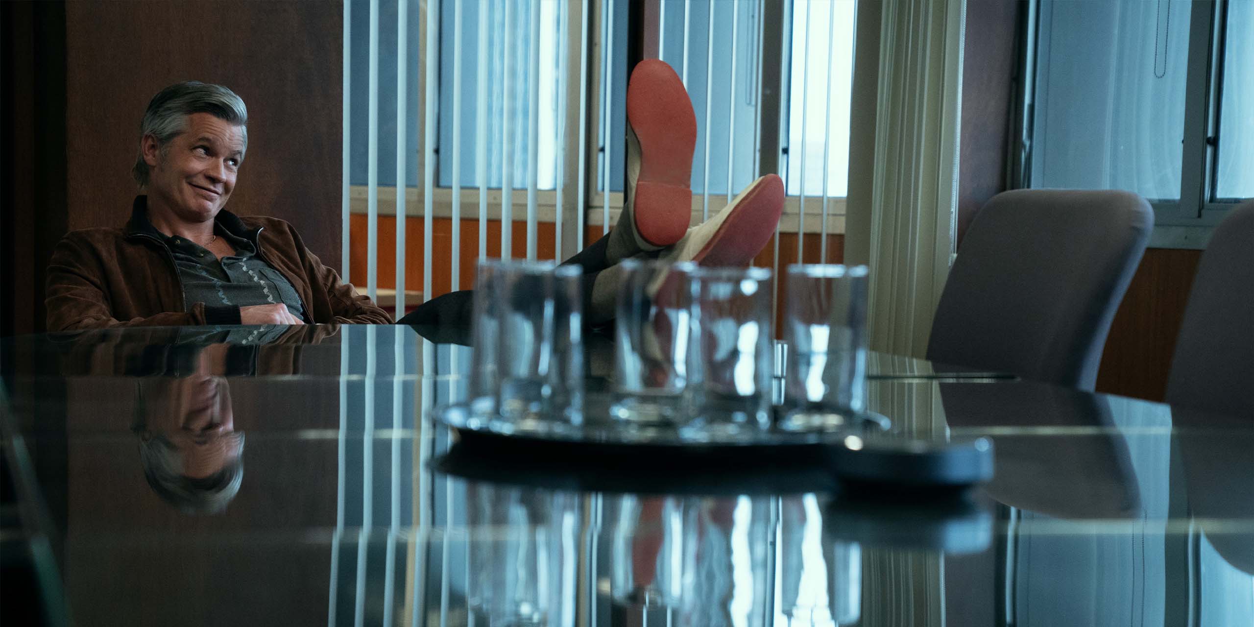 John Armstrong (Timothy Olyphant) dans une salle de réunion, les pieds sur la table, dans Lucky.