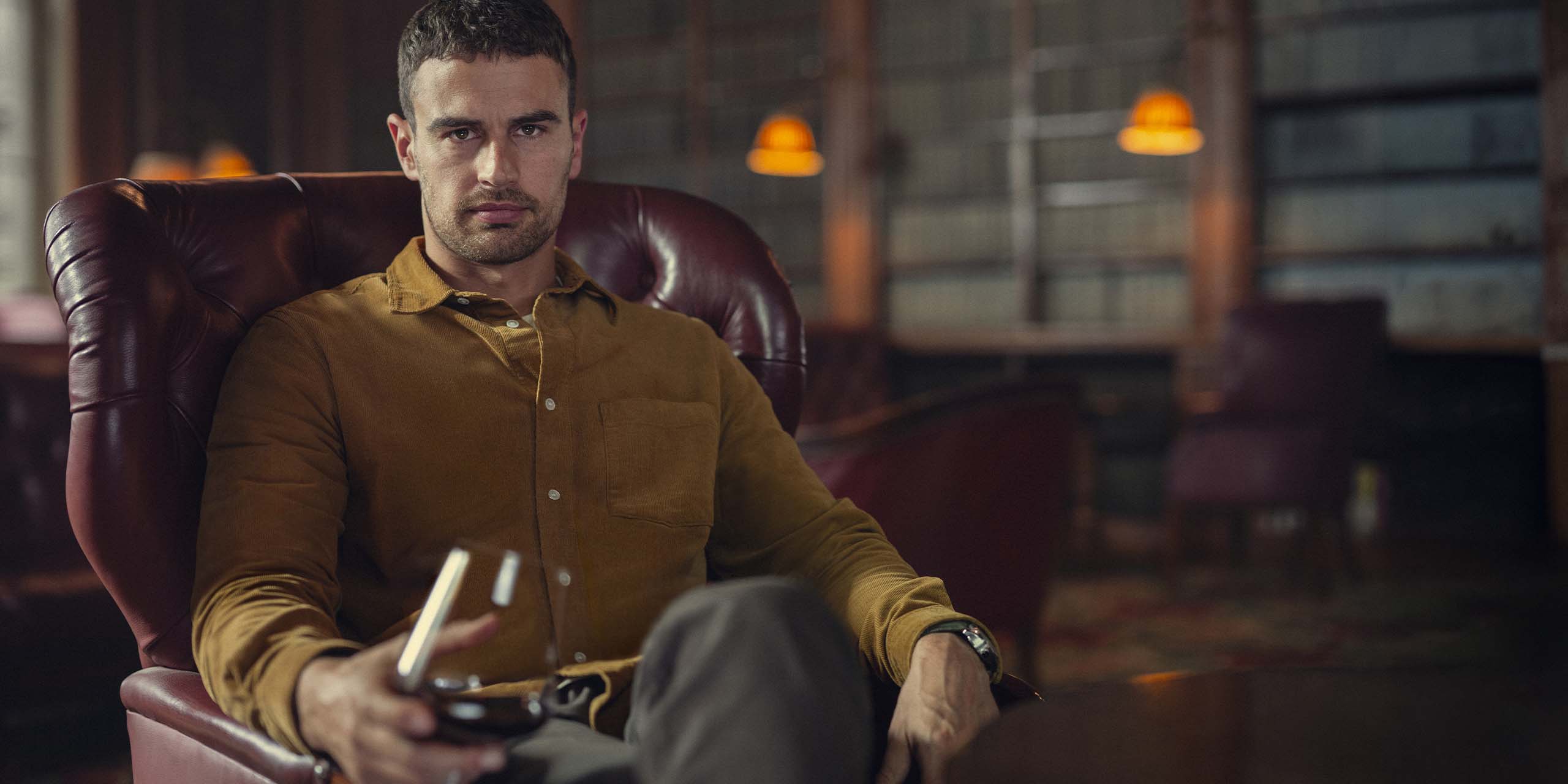 Eddie Horniman (Theo James) assis dans la bibliothèque avec un verre de vin, dans The Gentlemen.
